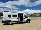 White Winnebago Solis camper van for RV rental, parked on gravel under cloudy sky. Side door open, revealing interior.
