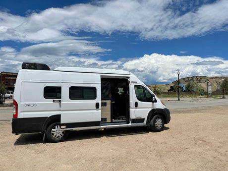 White Winnebago Solis camper van for RV rental, parked on gravel under cloudy sky. Side door open, revealing interior.