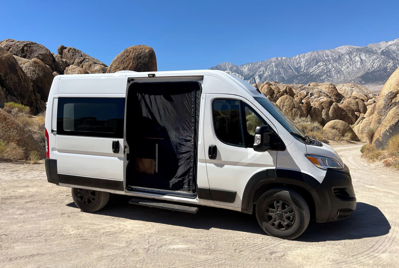 White campervan RV rental parked on a dirt road in a sunny desert with large rock formations and mountains. Side door open. — photo 12