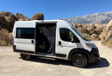 White campervan RV rental parked on a dirt road in a sunny desert with large rock formations and mountains. Side door open.