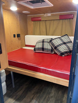 Cozy RV rental interior features a red mattress with plaid pillows, wood paneling, and an overhead vent, creating a functional sleeping area.