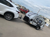 White SUV towing a small teardrop travel trailer, parked at an RV rental lot with larger campers.