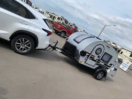 White SUV towing a small teardrop travel trailer, parked at an RV rental lot with larger campers.