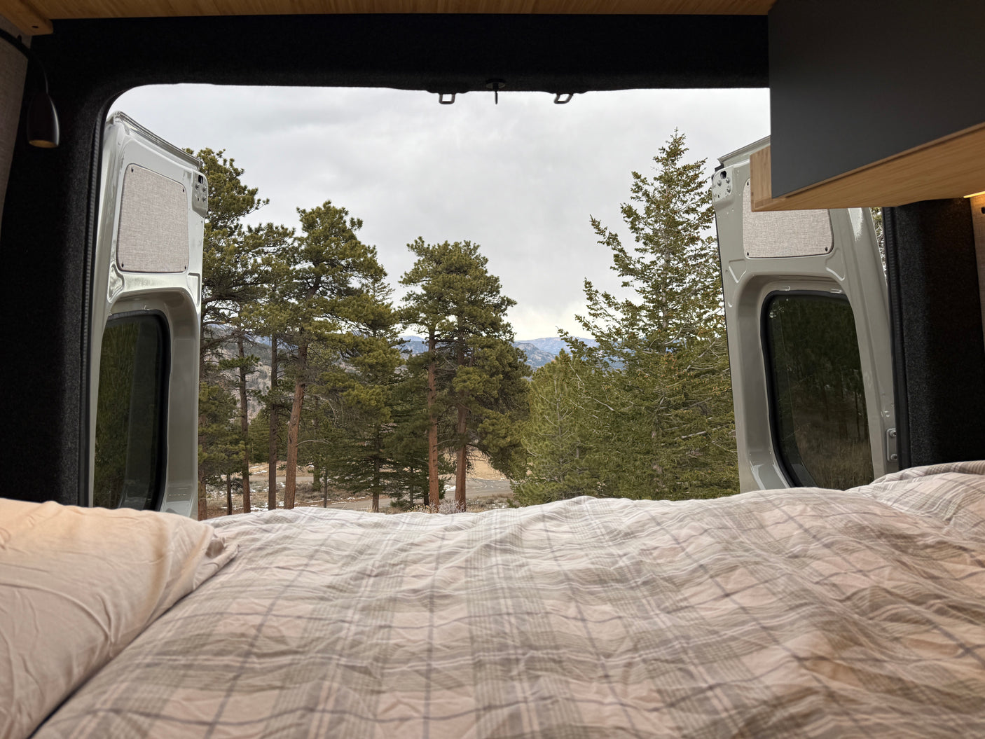 Enjoy nature from your RV rental! Cozy bed with plaid bedding looking out to a serene forest and mountains from a camper van.