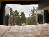 Enjoy nature from your RV rental! Cozy bed with plaid bedding looking out to a serene forest and mountains from a camper van.