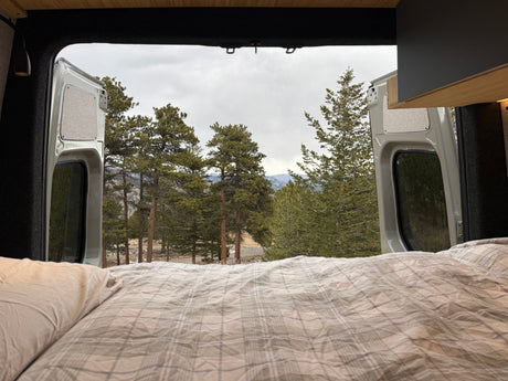 Enjoy nature from your RV rental! Cozy bed with plaid bedding looking out to a serene forest and mountains from a camper van.