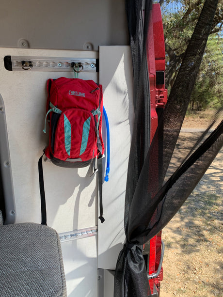 RV rental interior: A red Camelbak hangs on an E-track, with a black bug screen tied back, revealing trees outside.