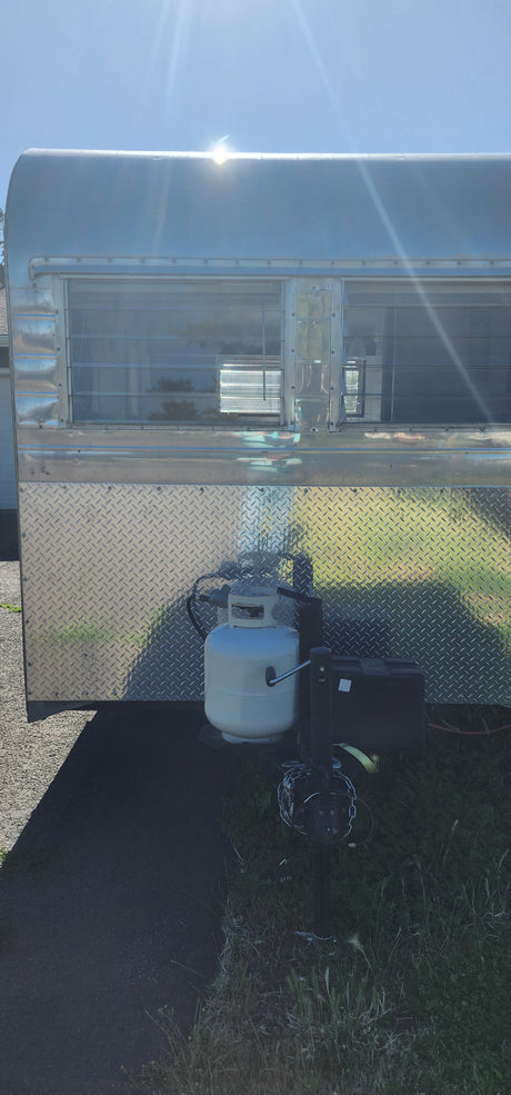 Front of a metallic RV rental travel trailer, featuring a white propane tank, battery, and hitch on a sunny day.