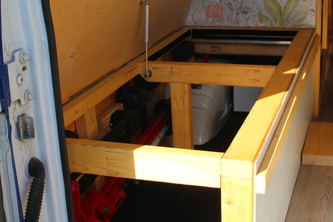 Inside an RV rental: a wooden bed frame with a hinged panel supported by a gas strut, revealing practical under-bed storage.