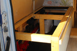 Inside an RV rental: a wooden bed frame with a hinged panel supported by a gas strut, revealing practical under-bed storage.