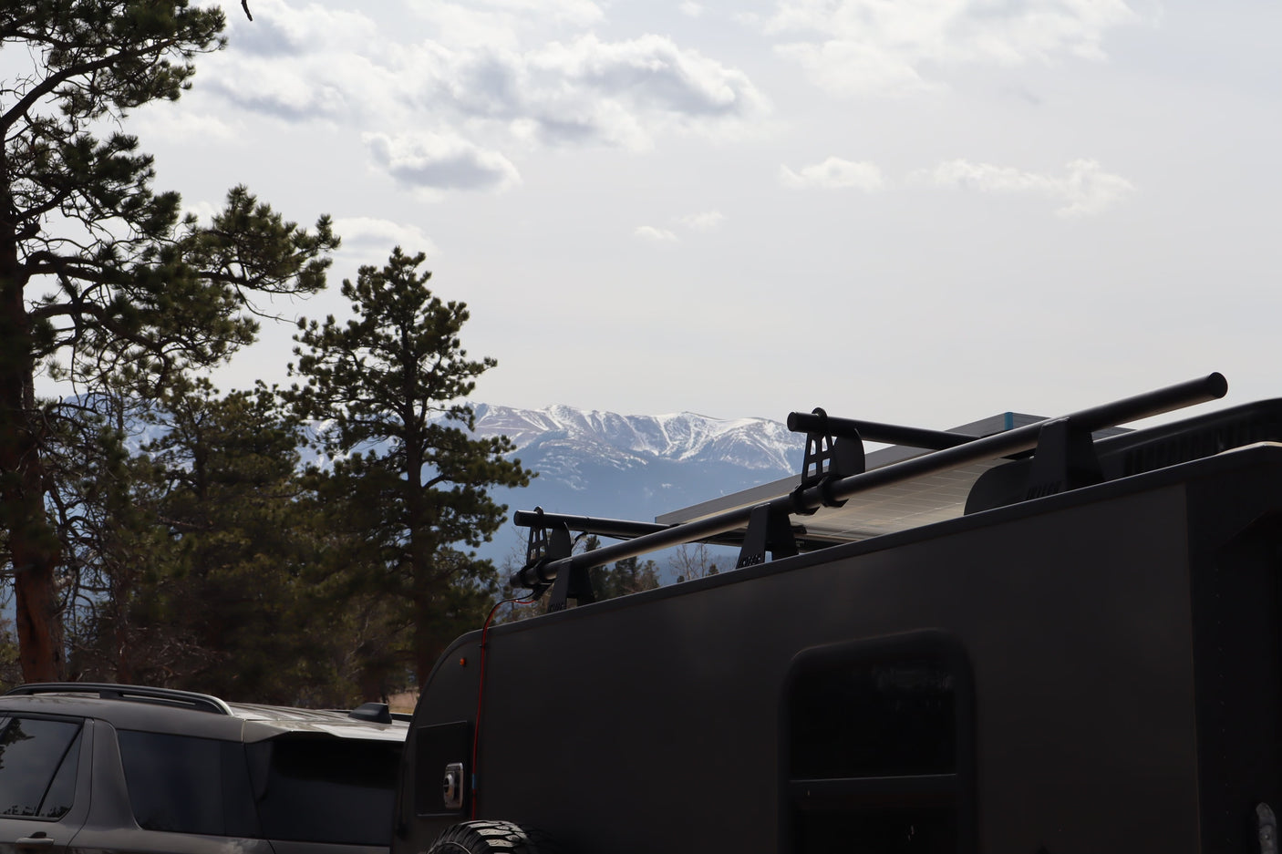 RV rental adventure: dark camper trailer with solar power, parked near pines with snowy mountains in the background.