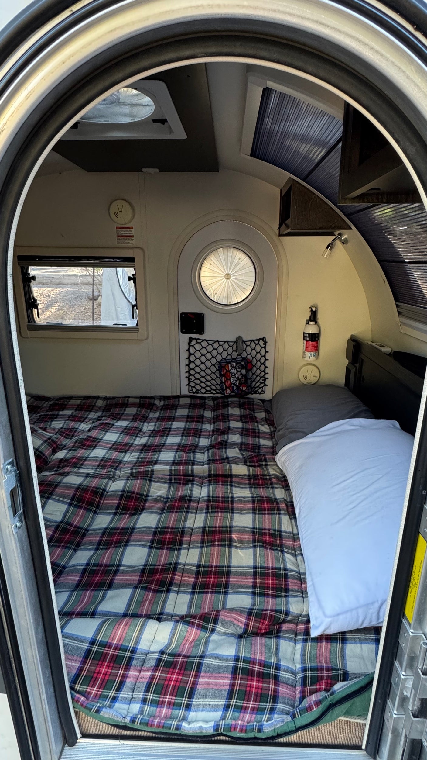 Interior view of an RV rental: a cozy camper bed with a plaid blanket, windows, and storage netting.
