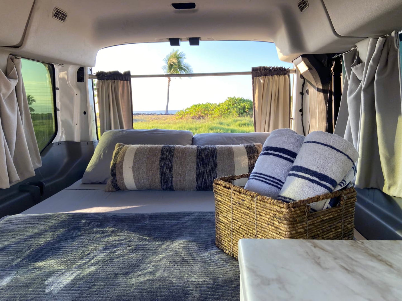 Inside a cozy RV rental van with a bed, plush blanket, fresh towels, and a beautiful tropical view out the back.