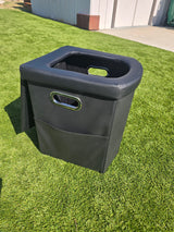 A black, portable RV rental toilet on green turf, featuring a U-shaped seat and side handles.
