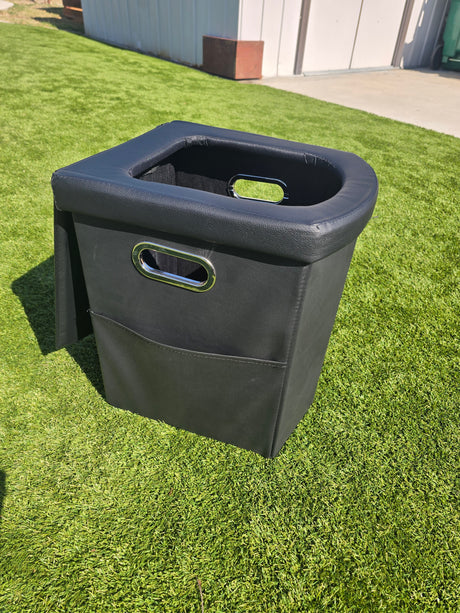A black, portable RV rental toilet on green turf, featuring a U-shaped seat and side handles.