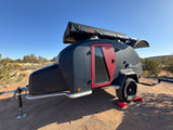 Ready for adventure: a rugged black teardrop RV rental with red accents and a rooftop tent, parked off-road in a sunny, dry landscape.