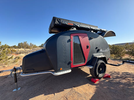 Ready for adventure: a rugged black teardrop RV rental with red accents and a rooftop tent, parked off-road in a sunny, dry landscape.
