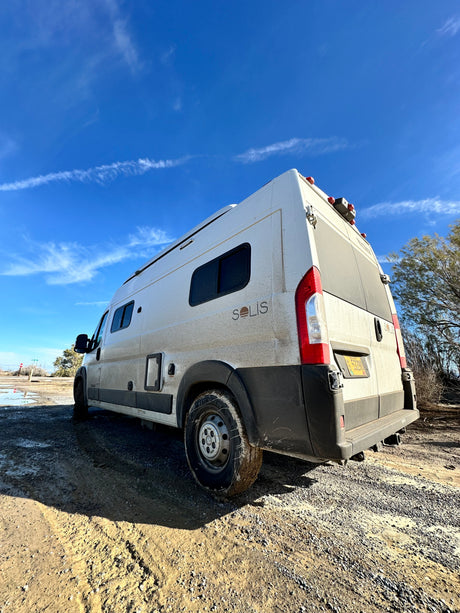 This dirty white Winnebago Solis RV rental stands on a muddy off-road trail under a clear blue sky, ready for adventure.