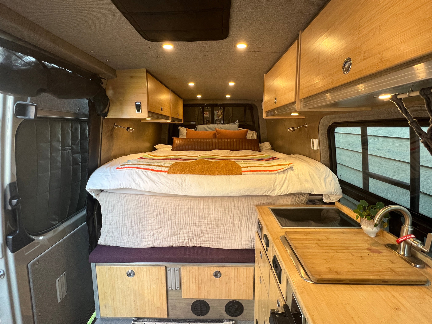 Inside a modern RV rental: a spacious bed, overhead storage, and kitchen area with a sink & cutting board.
