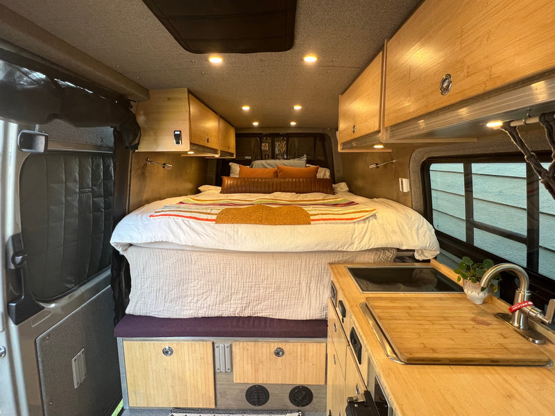 Inside a modern RV rental: a spacious bed, overhead storage, and kitchen area with a sink & cutting board. — photo 11