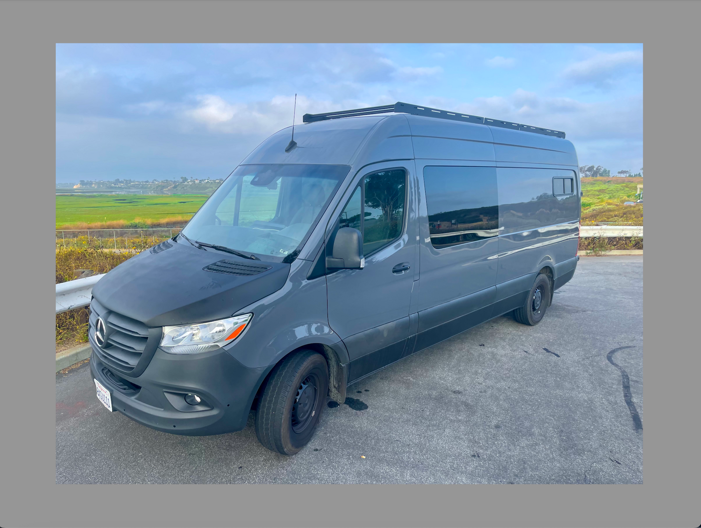 A modern gray Mercedes-Benz Sprinter van, likely an RV rental, with a roof rack, parked overlooking green fields. — photo 6