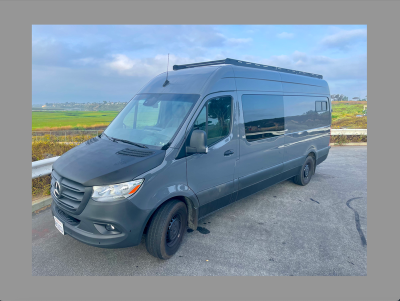 A modern gray Mercedes-Benz Sprinter van, likely an RV rental, with a roof rack, parked overlooking green fields. — photo 6