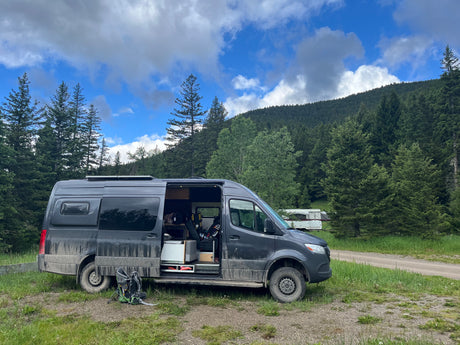 Scenic RV rental camper van parked with open door at a forested mountain campsite. Dirty van suggests outdoor adventure.