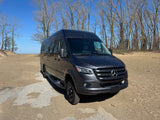 Explore with this dark gray Mercedes Sprinter RV rental. Parked on a sandy path near water, with bare trees under a blue sky.