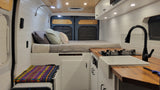 Bright & modern RV rental interior: cozy bed, compact kitchen with wood counter, and white storage.