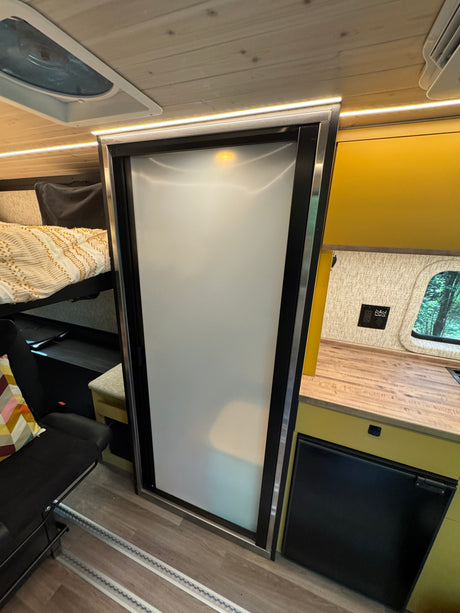 Modern RV rental interior: cozy bunk bed, frosted glass door (likely bath), yellow kitchen, wood ceiling, fridge, bright lighting.