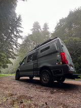 Rugged RV rental van on a wet, misty forest road. Tall trees frame this adventure scene.