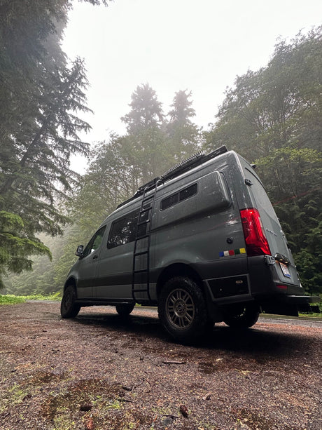 Rugged RV rental van on a wet, misty forest road. Tall trees frame this adventure scene.