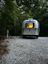 A sleek Airstream rv rental glows softly inside, parked on a gravel campsite amidst dense forest, hooked up for a cozy stay.