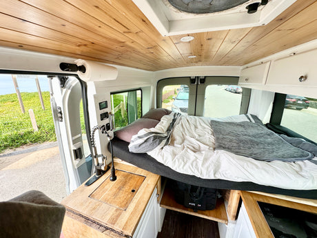 Inside a cozy RV rental camper van, revealing a bed, wooden ceiling, and kitchenette. Outside: green landscape & road.
