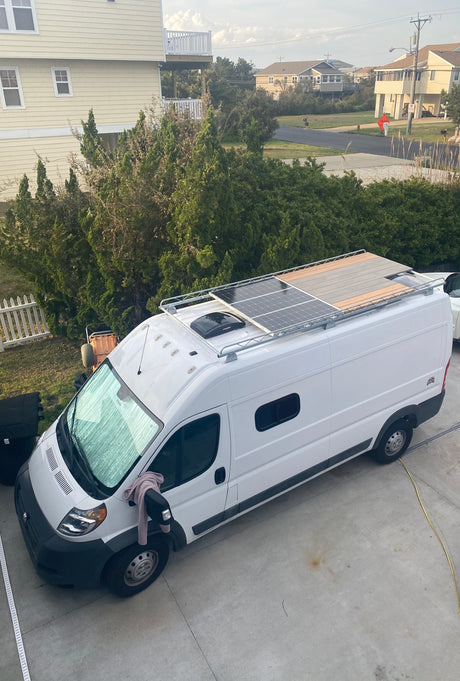 White **RV rental** campervan with solar panels on its roof rack, parked on a driveway beside a house.