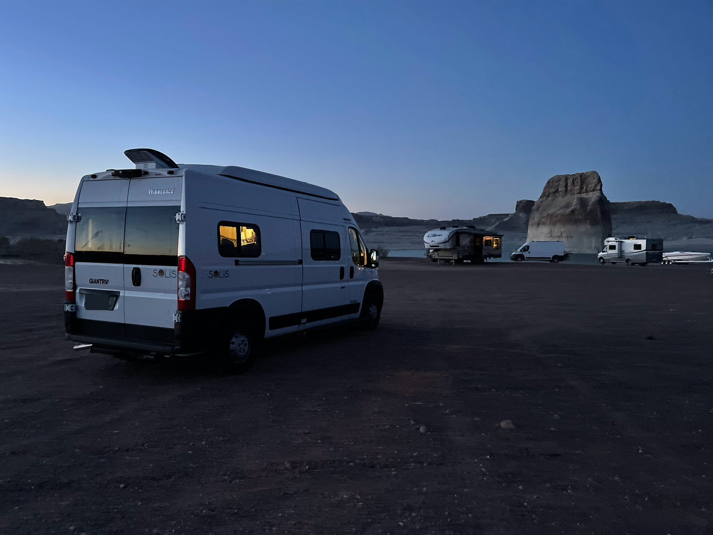 White Winnebago Solis RV rental parked at a dark campsite with other RVs, boat, and rock formations in distance.