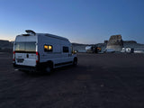 White Winnebago Solis RV rental parked at a dark campsite with other RVs, boat, and rock formations in distance.