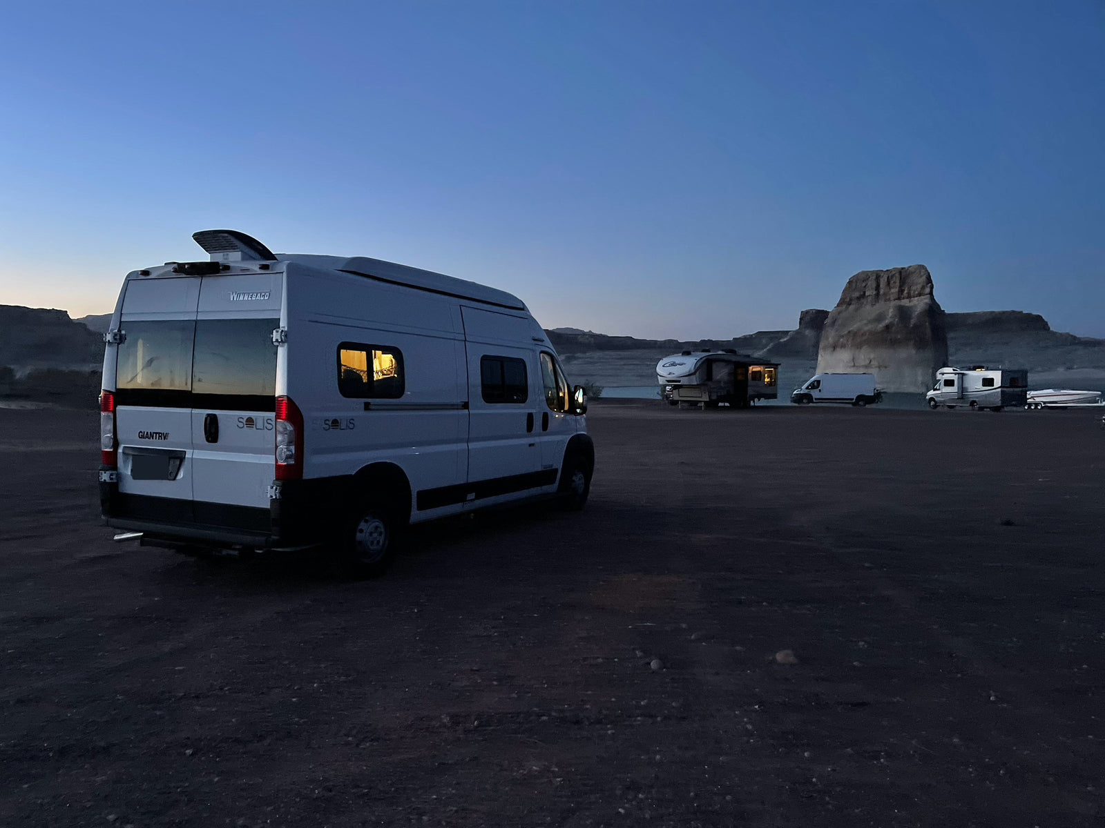 White Winnebago Solis RV rental parked at a dark campsite with other RVs, boat, and rock formations in distance.