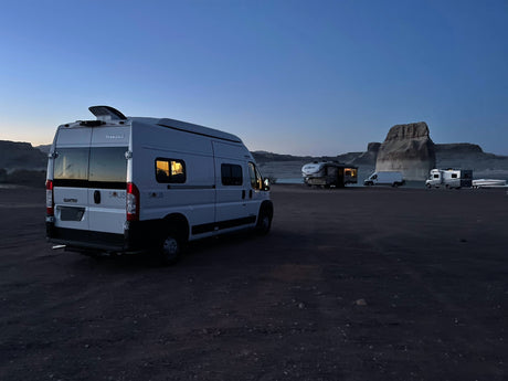 White Winnebago Solis RV rental parked at a dark campsite with other RVs, boat, and rock formations in distance.