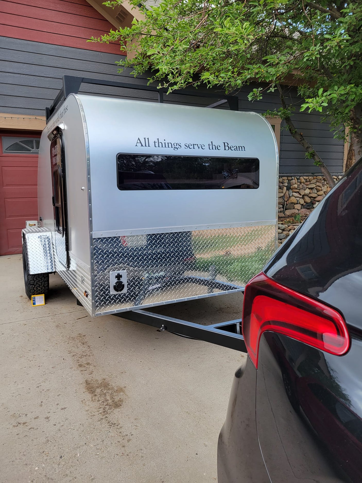 A silver teardrop camper, ideal for RV rental, hitched to a dark car in a driveway beside a house and tree.