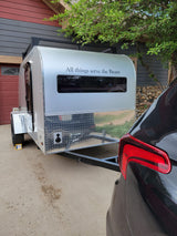 A silver teardrop camper, ideal for RV rental, hitched to a dark car in a driveway beside a house and tree.