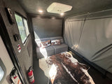 RV rental interior: cozy living space with modular grey sofa, cowhide rug, window, and ceiling AC unit. Fire extinguisher visible.