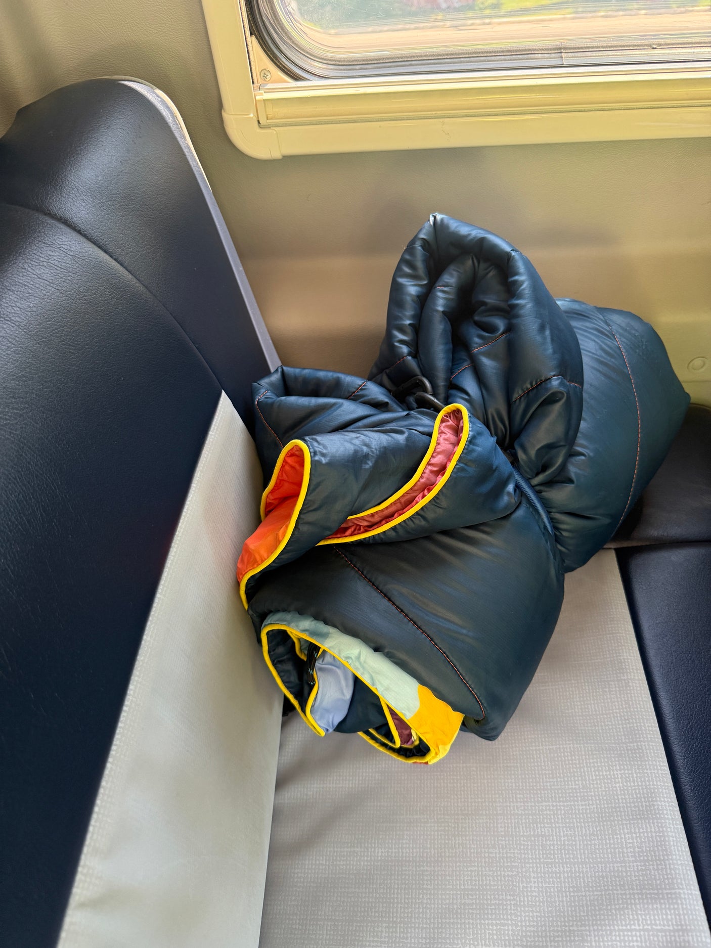 Inside an RV rental: a dark blue sleeping bag, with bright orange and yellow trim, rests on patterned grey seating beside a window.