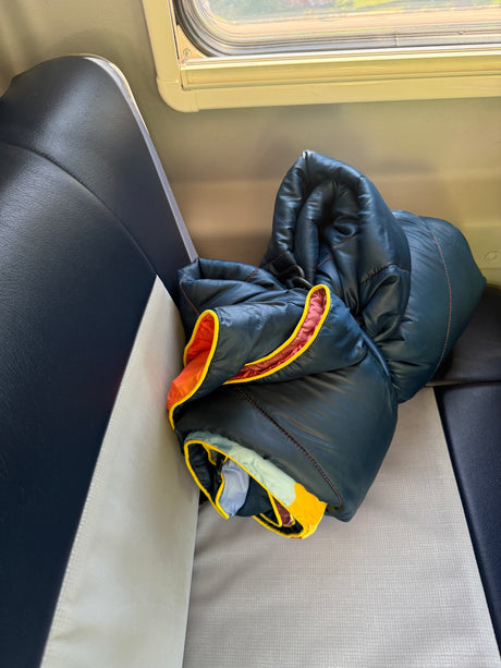 Inside an RV rental: a dark blue sleeping bag, with bright orange and yellow trim, rests on patterned grey seating beside a window.