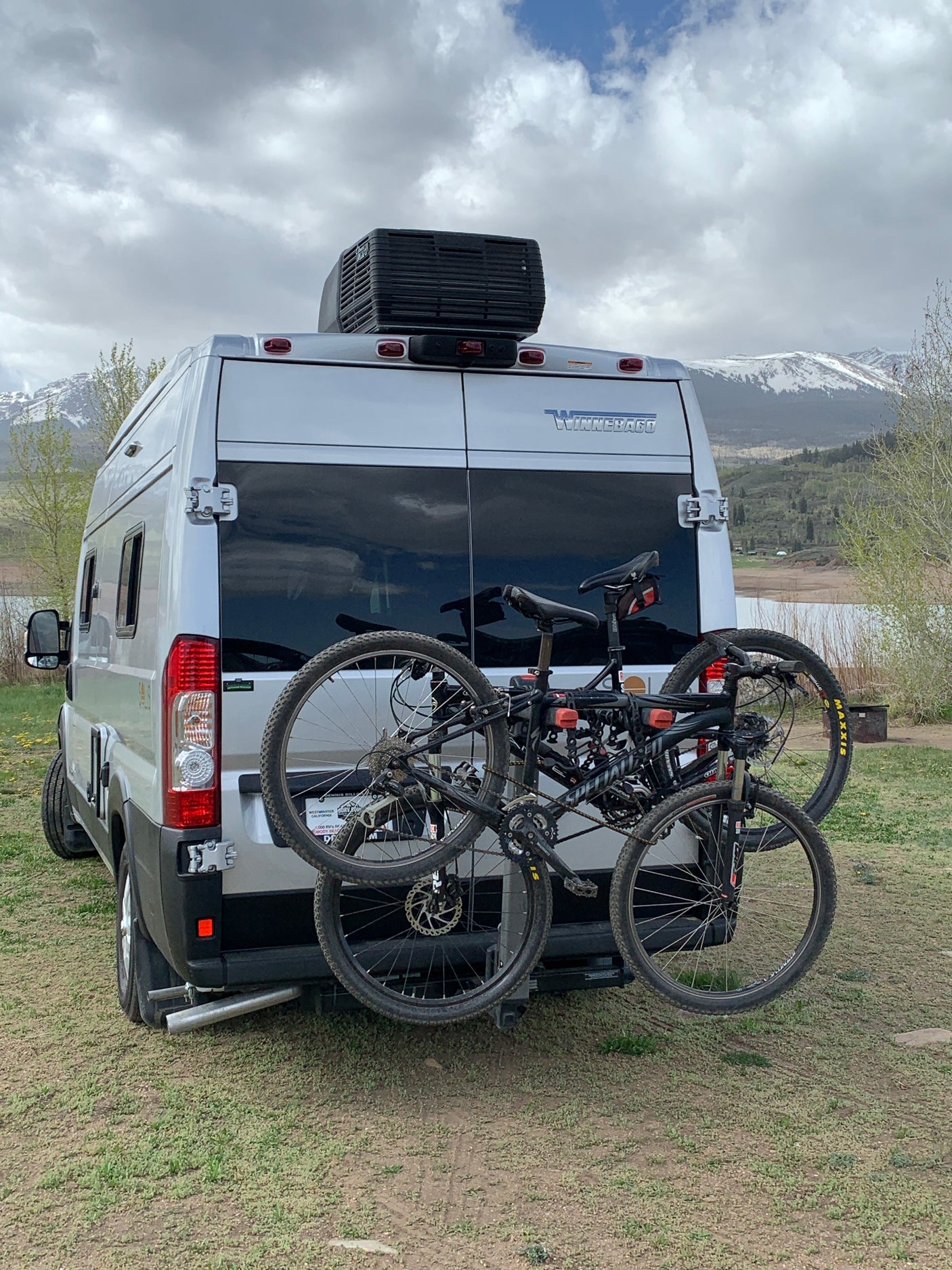 Silver RV rental with mountain bikes parked by a lake, snowy mountains in the background. Ready for adventure!