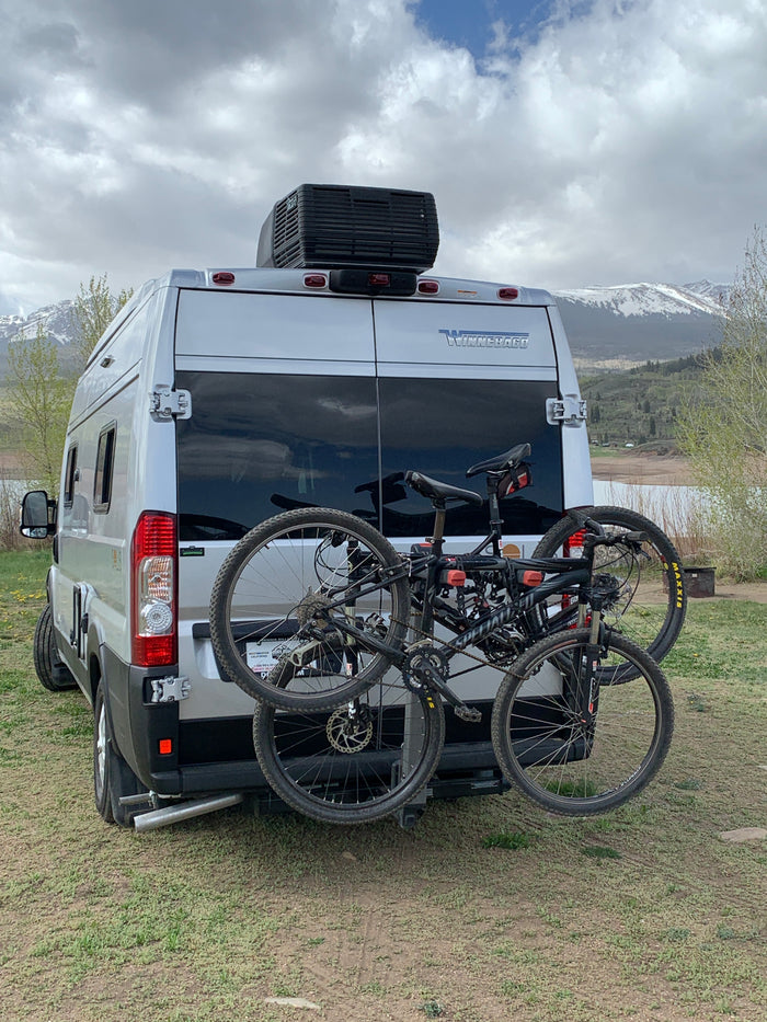 Silver RV rental with mountain bikes parked by a lake, snowy mountains in the background. Ready for adventure!