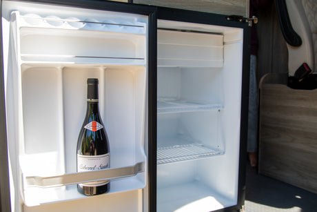 Inside an RV rental, a compact fridge is open, showing a Cabernet-Syrah wine bottle in its door and empty shelves.