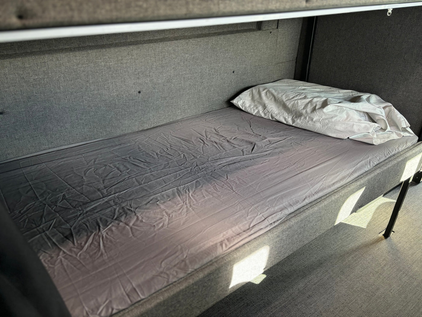 Inside an RV rental: a cozy lower bunk bed with grey fabric walls, a wrinkled grey sheet, and a white pillow, illuminated by sunlight. — photo 15