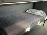 Inside an RV rental: a cozy lower bunk bed with grey fabric walls, a wrinkled grey sheet, and a white pillow, illuminated by sunlight.