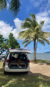 A white SUV RV rental, trunk open revealing a bed, parked by a tropical ocean under palm trees and blue sky.